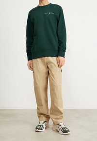 Mörkgrön sweatshirt med rund hals och tryckt "GANT 1949"-logotyp, matchad med ljusbeiga cargobyxor och sneakers med gröna och vita accenter.