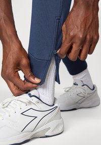 Hände ziehen die Reißverschlussöffnung der navyblauen Sporthose über der weißen Socke und dem weißen Sneaker mit dunkelblauen Akzenten auf hellem Hintergrund auf.