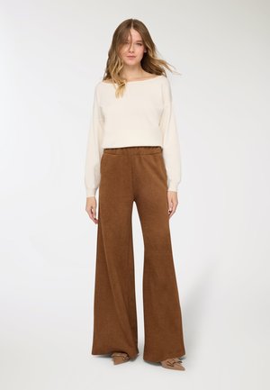 Top avorio a maniche lunghe con scollo ampio; pantaloni marroni a gamba larga con tessuto strutturato. Calzature di colore marrone chiaro con dettagli decorativi.