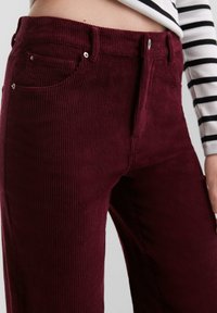 Bordeaux corduroy broek met verticale ribbels, vijfzakontwerp en metalen hardware accenten. De stof heeft een zachte, gestructureerde afwerking.