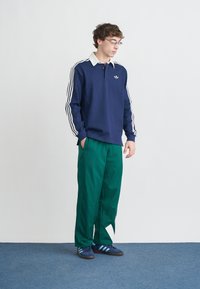 Μπλε σκούρο πόλο με μακριά μανίκια και λευκό γιακά, με λευκές ρίγες στα μανίκια. Συνδυάζεται με πράσινο παντελόνι και μπλε αθλητικά παπούτσια με τρεις ρίγες.