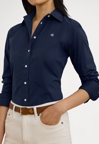 Temno modra srajca z gumbi, ovratnikom, dolgimi rokavi in majhnim vezenim logotipom. Kombinirana s kremasto barvnimi hlačami in rjavim pasom.