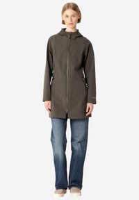 Chaqueta impermeable de tela suave en gris oscuro con capucha, cremallera frontal y cordones ajustables a los lados. Combinada con jeans acampanados de color azul claro.