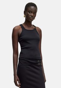 Zwarte geribbelde tanktop met een gouden accent op de schouder. Het ontwerp heeft een aangesloten vorm en dikke bandjes, gecombineerd met een zwarte rok.