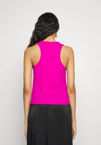 Vrouw met lang, golvend zwart haar, gekleed in een mouwloos felroze topje en losse zwarte broek, staand tegen een effen lichte achtergrond.