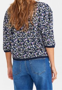 Frau mit langen braunen Haaren, die ein marineblusiges Oberteil mit pinken und weißen Akzenten sowie blaue Jeans trägt, von hinten gezeigt.