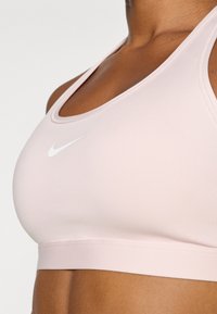 Svetlo roza športni modrček z širokim podaljškom, z belim Nike logotipom spredaj. Gladek material, oblikovanje s športnim hrbtom in brezšivne robove.