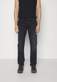Jeans in denim blu scuro con fitte dritta, design a cinque tasche, leggero sbiadimento e dettagli di cucitura sottili. Indossati con scarpe da trekking nere.