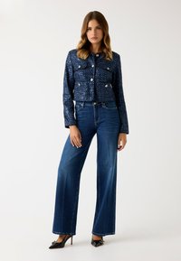 Donna che indossa un giaccone blu navy texturizzato con bottoni, jeans a vita alta e gamba larga di un blu scuro, e tacchi neri a punta con dettaglio a fibbia.