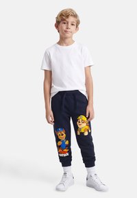 Αγόρι με ξανθά μαλλιά που στέκεται φορώντας λευκό μπλουζάκι, σκούρες μπλε φόρμες με χαρακτήρες από το Paw Patrol, λευκές κάλτσες και λευκά αθλητικά παπούτσια σε λευκό φόντο.