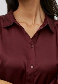 Burgundy satin blouse with a collared neckline, button closure, and subtle sheen. Features short sleeves and a smooth texture.
