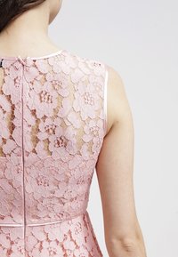 Růžové krajkové šaty s květinovými vzory, průsvitným zadním dílem a diskrétním zapínáním na zip, s saténovým lemem kolem výstřihu a okrajů rukávů.