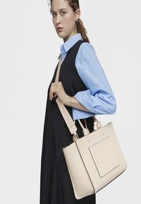 Beżowa torebka z wegańskiej skóry z podwójnymi uchwytami i odpinanym paskiem. Charakteryzuje się wytłoczonym logo oraz usystematyzowanym prostokątnym kształtem.