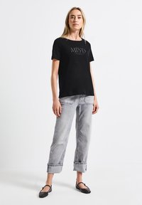 T-shirt noir à manches courtes avec un graphique à l'avant portant le mot "MIND" en gris clair. Associé à un jean gris clair retroussé et des ballerines noires.