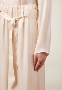Lichtbeige jurk met een aangesloten elastische tailleband en een geknoopt stoffen riem. Lange, losse mouwen met een gladde textuur.