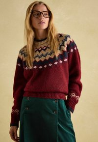 Roter Wollpullover mit beigem und blauem Muster im Schulterbereich, kombiniert mit einem grünen Cordrock mit Knopfakzenten. Strukturiertes Material.