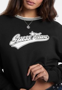 Felpa nera con un grande logo bianco "Guess Jeans", collo a righe e texture morbida. Il modello tiene il tessuto vicino all'orlo.