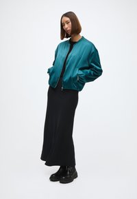 Türkiz selyem bomber dzseki cipzárral, fekete maxi ruhára és fekete vastag bokacsizmára viselve, oldalsó zsebekkel és elasztikus alsó résszel.