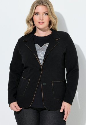 Blond vrouw die een zwarte blazer met bruine stiksels draagt over een zwart shirt met een zilveren pailletten hart, staand tegen een witte muur.