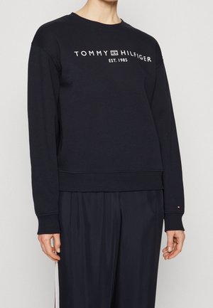Persoon draagt een zwarte Tommy Hilfiger sweater met witte logotekst "TOMMY HILFIGER EST. 1985" en zwarte broek met witte zijstreep.