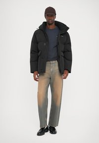 Black puffer jacket with a hood, matte finish, and patch details, paired with faded wide-leg jeans and black shoes. Dark cap completes the look.