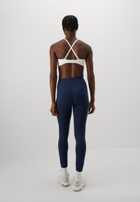 Person som står med ryggen mot kameran, iklädd en vit sport-bh med korsande axelremmar, marinblå högmidjade leggings, vita strumpor och beige sneakers.