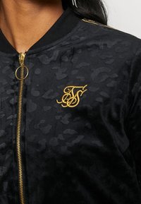 Giubbotto bomber nero con stampa leopardata testurizzata, caratterizzato da una zip dorata e un logo ricamato sul petto. Materiale simile al velluto.