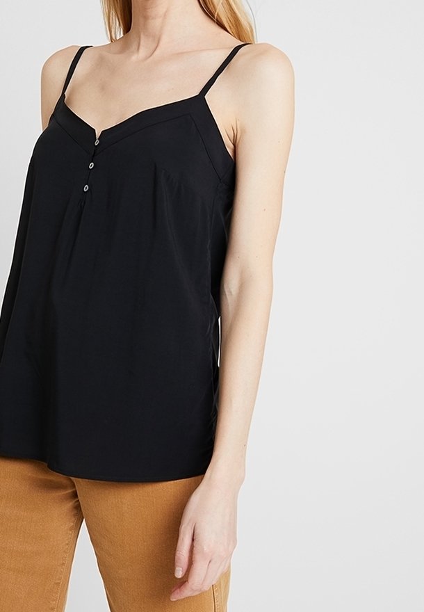 Kvinna klädd i en svart ärmlös topp med knappar och kamelfärgade byxor, stående mot en plain ljus bakgrund.