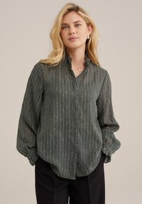 Áttetsző zöld blúz, texturált függőleges csíkokkal, fodros gallérral és hosszú ujjakkal, elegáns fekete nadrággal viselve.