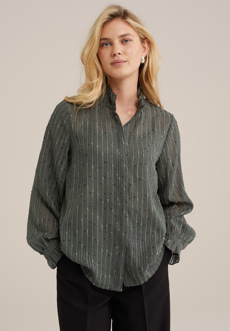 Áttetsző zöld blúz, texturált függőleges csíkokkal, fodros gallérral és hosszú ujjakkal, elegáns fekete nadrággal viselve.