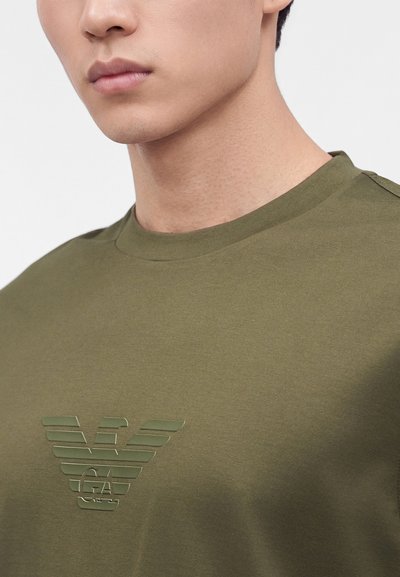 Νεαρός άνδρας με ελιάς πράσινο T-shirt με ανάγλυφο γυαλιστερό λογότυπο αετού στο στήθος, κοντινό πλάνο στον λαιμό και το άνω μέρος του κορμού.