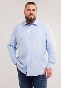 Mann mit Bart, der ein hellblaues Hemd und dunkle Jeans trägt, steht mit einer Hand in der Tasche vor einem einfarbigen Hintergrund.