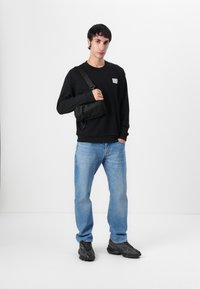 Felpa nera con una piccola toppa con logo, jeans azzurri chiari e sneakers nere. Una borsa a tracolla nera è indossata sulla spalla.