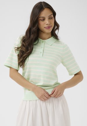 Žena s dlhými tmavými vlasmi, oblečená v svetlozelenom a bielo pruhovanom krátkym rukávom polo tričku a bielej sukni, upravuje lem trička.