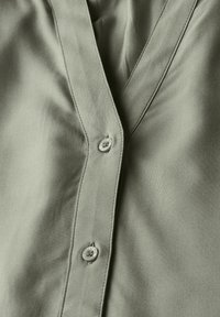 Camicia verde oliva con colletto a V profondo, chiusura con un bottone e texture di tessuto liscia, caratterizzata da sottili dettagli di cucitura.
