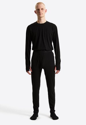 Junge Person steht, trägt ein eng anliegendes schwarzes Langarmshirt und passende schwarze Hose mit schwarzen Socken vor weißem Hintergrund.