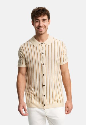 Homme souriant portant une chemise en tricot à manches courtes, beige et marron à rayures, boutonnée, avec un pantalon blanc sur un fond uni.