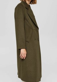 Kobieta w długim oliwkowozielonym wełnianym płaszczu dwurzędowym z szerokimi klapami i bocznymi kieszeniami, pokazana z boku.