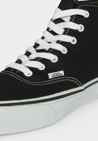 Μαύρο ψηλό αθλητικό παπούτσι Vans με λευκά κορδόνια, λευκή ραφή και λευκή λαστιχένια σόλα, που δείχνει πλαϊνό ετικέτα με λογότυπο.