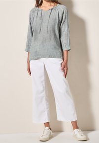 Femme portant un chemisier à manches longues rayé gris, un pantalon large blanc et des baskets blanches, debout contre un mur beige.