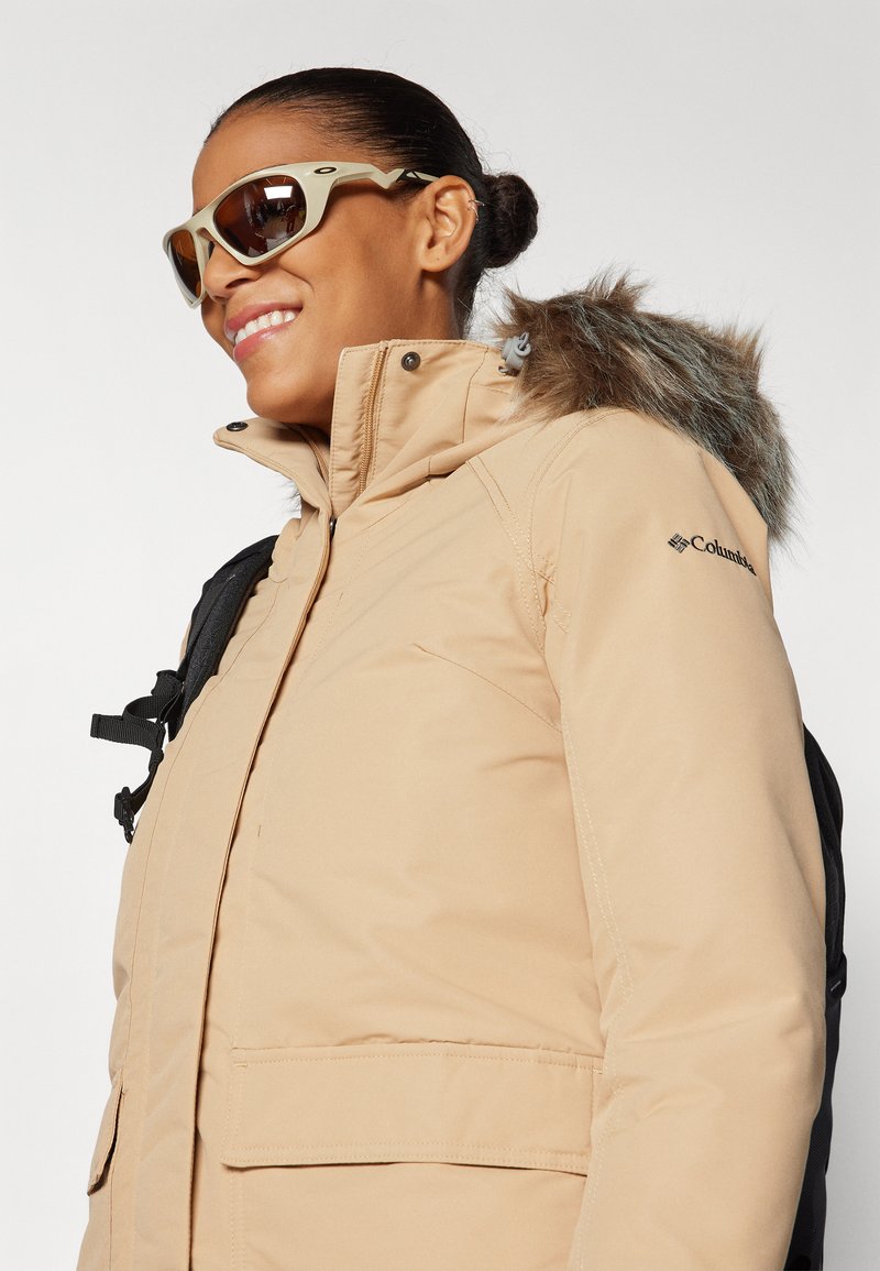 Veste isolante beige avec capuche amovible en fourrure, équipée d'une fermeture éclair et de poches. Accompagnée de lunettes de soleil et d'un sac à dos noir.