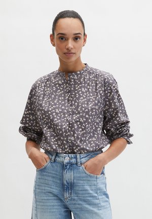 Vrouw met donker haar in een knot, gekleed in een donker gebloesd blouse met lange mouwen, ingestopt in lichtblauwe, hooggesneden spijkerbroek, met handen in de zakken.