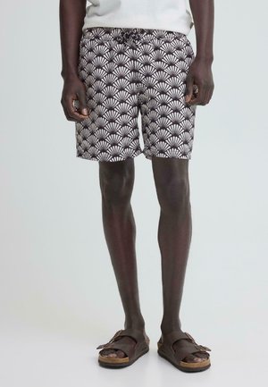 Man draagt zwart-witte korte broek met patroon en bruine sandalen met gesp, staand tegen een effen lichte achtergrond.