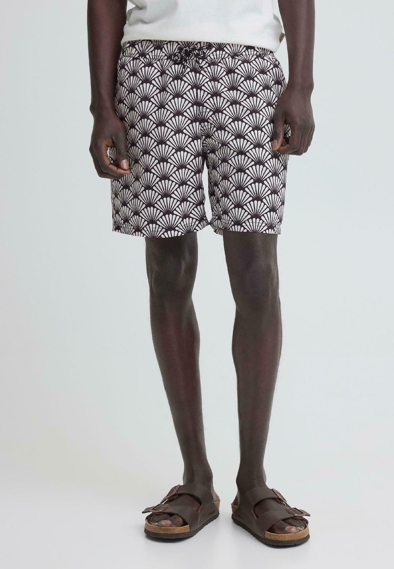 Άνδρας που φοράει μαύρο-άσπρο σορτς με σχέδια και καφέ σανδάλια με αγκράφα, στέκεται μπροστά σε απλό ανοιχτόχρωμο φόντο.