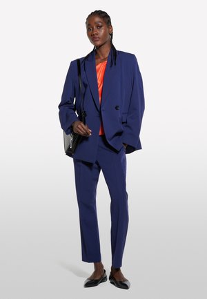 Completo blu navy con giacca doppiopetto, blusa arancione sotto e pantaloni sartoriali. Ballerine nere e una borsa a tracolla nera completano il look.