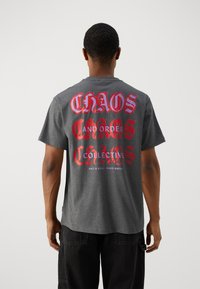 Pilkas trumpomis rankovėmis marškinėliai su dideliu raudonos ir purpurnos spalvos grafiniu tekstu ant nugaros, kuriame parašyta "CHAOS AND ORDER COLLECTIVE."