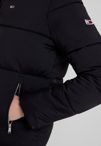 Svart puffjacka med dragkedja, vadderad design och ribbade muddar. Märkbar logotyp patch på vänster ärm.