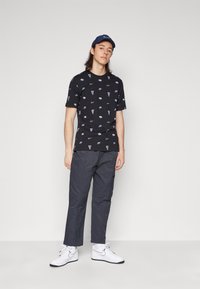 Camiseta negra con pequeños logos morados de Nike, combinada con pantalones grises y zapatillas blancas de Nike. El modelo lleva una gorra de béisbol azul marino.