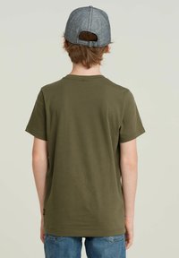 Oliven grøn bomulds t-shirt med korte ærmer, rund halsudskæring og et mærke nær kanten. Grå denim kasket båret baglæns.