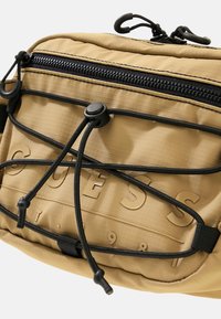 Beige Nylon-Crossbody-Tasche mit strukturiertem Oberflächen, schwarzem Reißverschluss und elastischen Kordeldetails. Verfügt über ein erhabenes Logo und einen verstellbaren Riemen.